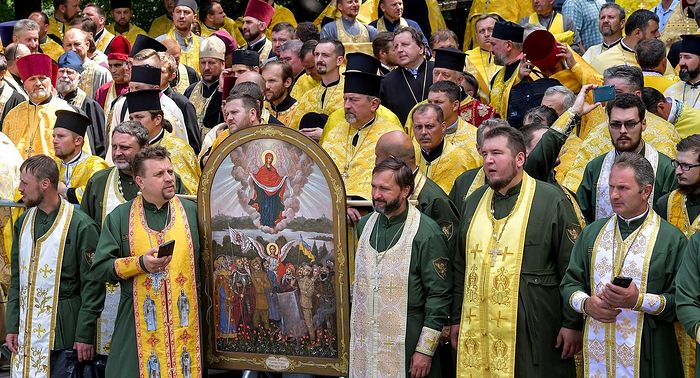 Schismatic “chaplains” in cassocks with military style patches carry an “icon” depicting various Ukrainian soldiers, including one armed with a Kalashnikov rifle and a Ukrainian flag.